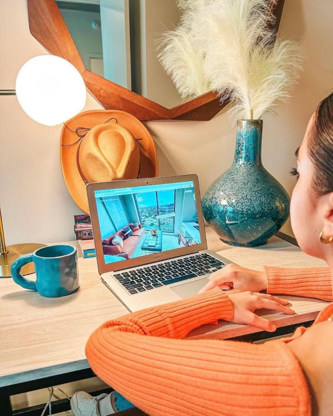 Rev Student Living Apartments Woman in an orange top uses a laptop at a desk with a mug, lamp, vase, and hat in a cozy, modern room.