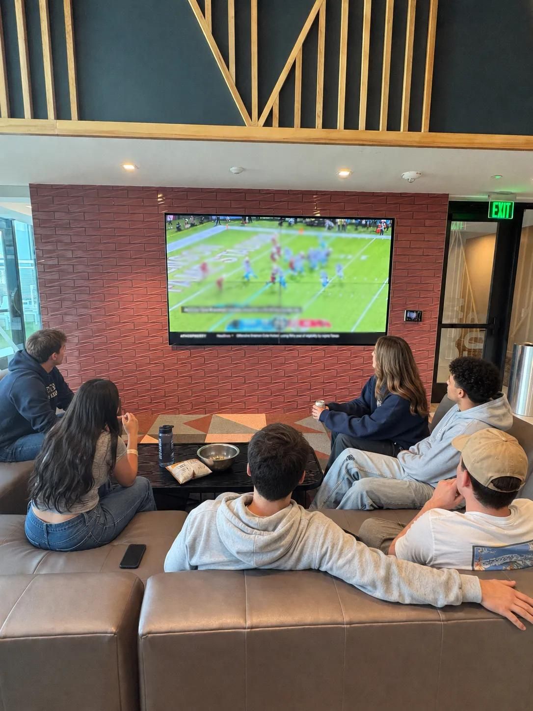 Rev Student Living Apartments Five people sit on couches in a modern living room, watching a football game on a wall-mounted TV. Where to watch aggie football.