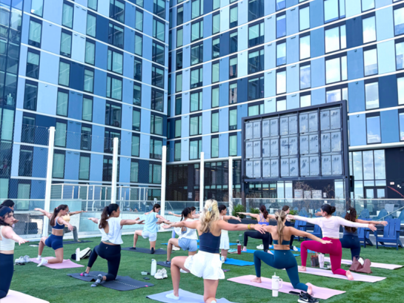Rev Student Living Apartments A group of women in activewear performing yoga on an outdoor turf terrace in front of a modern glass building.
