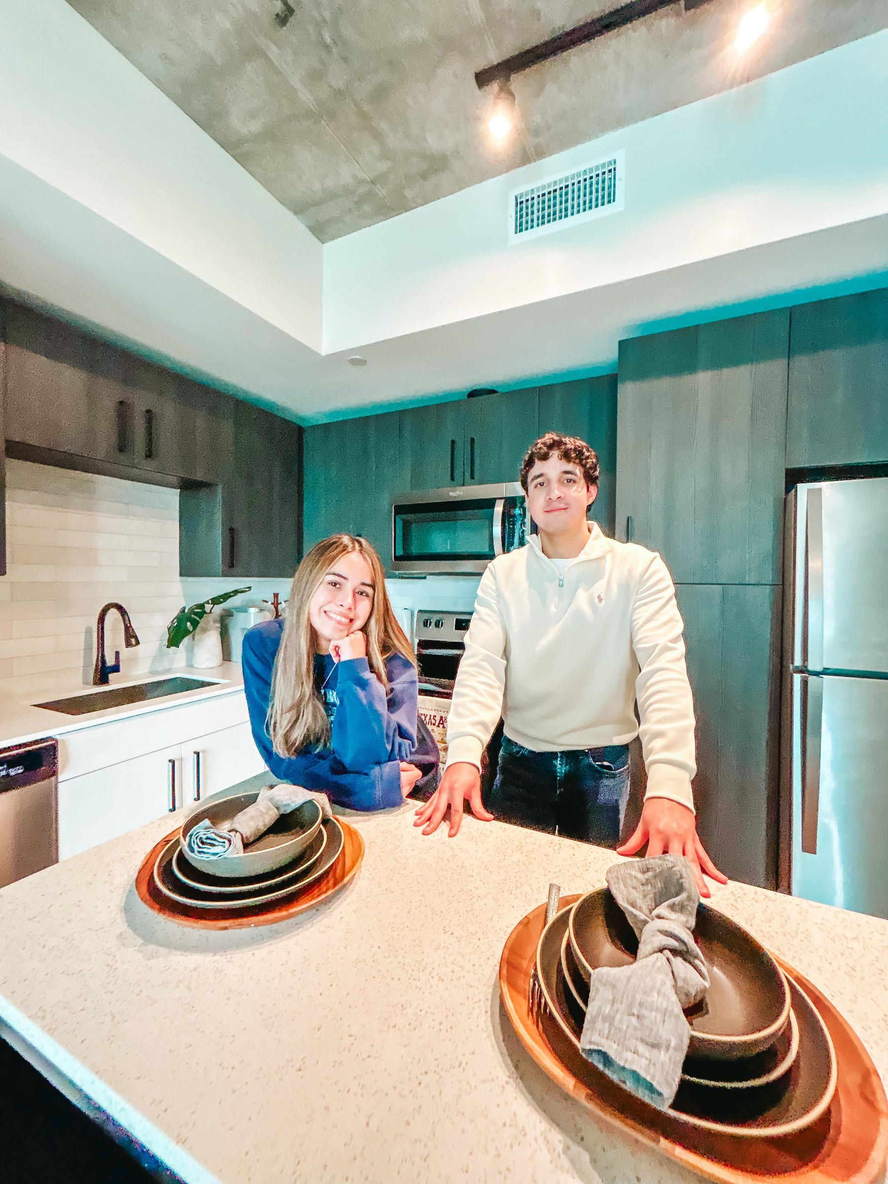 Rev Student Living Apartments Two people stand and smile in a modern kitchen behind a counter set with plates, napkins, and silverware.