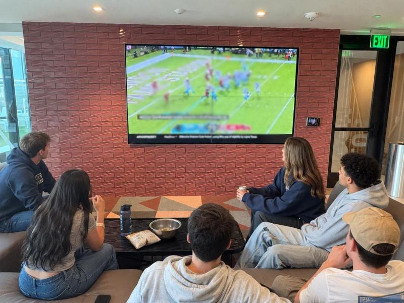 Rev Student Living Apartments Five people sit on couches in a modern living room, watching a football game on a wall-mounted TV. Where to watch aggie football.