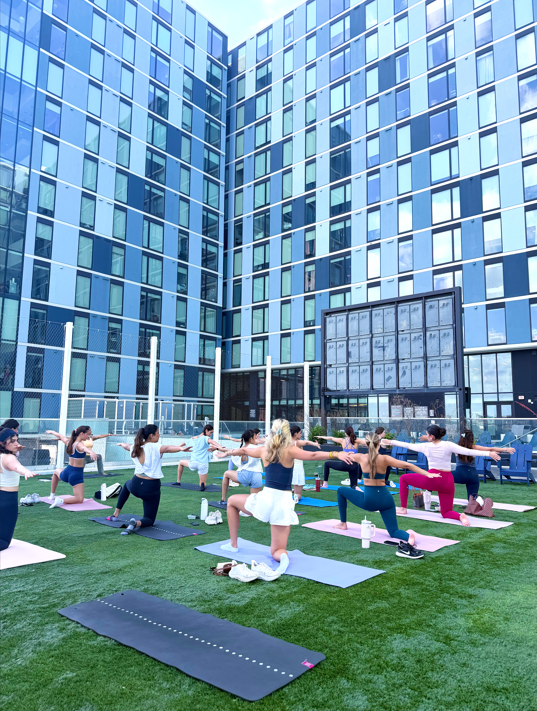 Rev Student Living Apartments A group of women in activewear performing yoga on an outdoor turf terrace in front of a modern glass building.