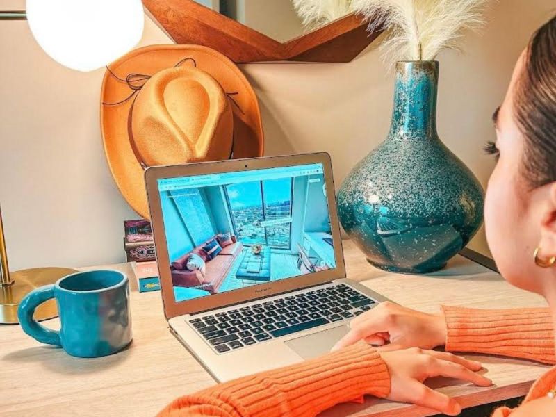 Rev Student Living Apartments Woman in an orange top uses a laptop at a desk with a mug, lamp, vase, and hat in a cozy, modern room.
