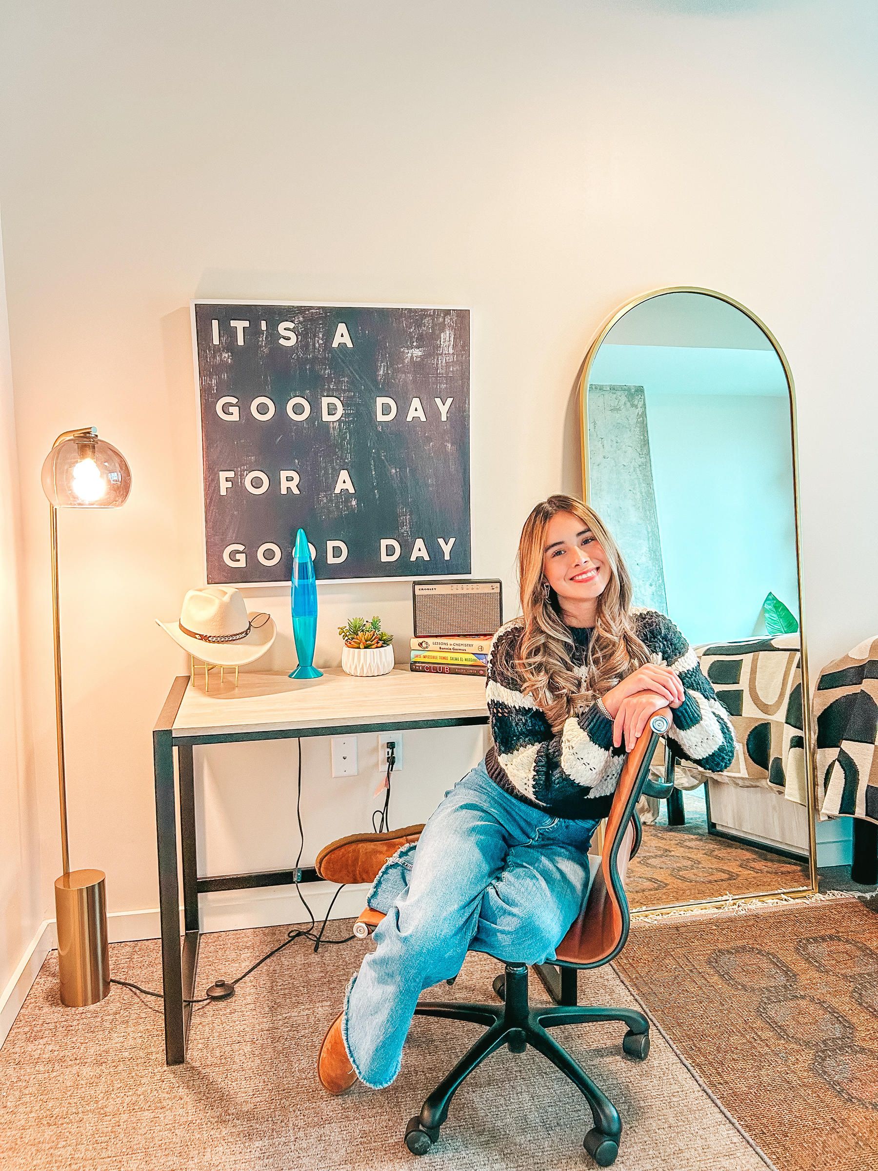 Rev Student Living Apartments Woman sitting on chair in cozy home office, smiling near desk with books, lamp, plants, and a sign that says “It’s a good day for a good day.”.