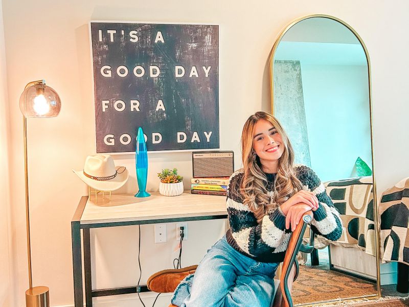 Rev Student Living Apartments Woman sitting on chair in cozy home office, smiling near desk with books, lamp, plants, and a sign that says “It’s a good day for a good day.”.