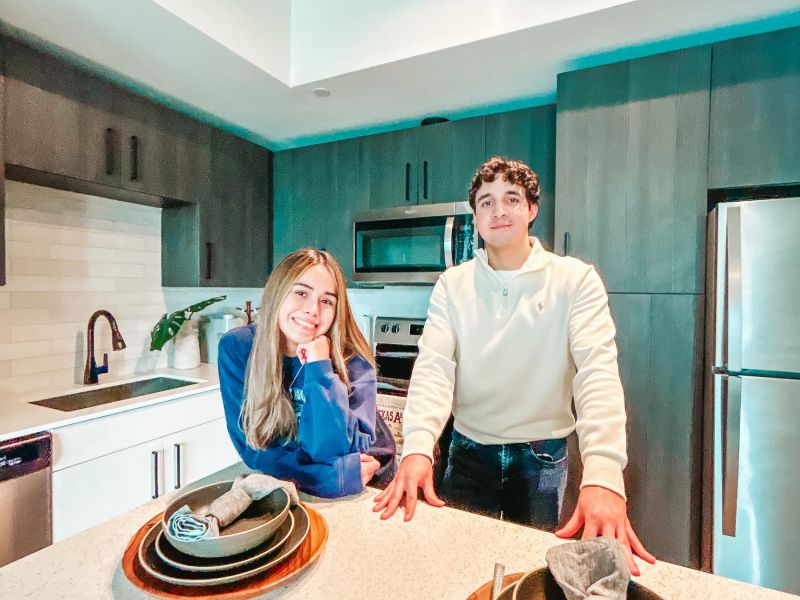 Rev Student Living Apartments Two people stand and smile in a modern kitchen behind a counter set with plates, napkins, and silverware.